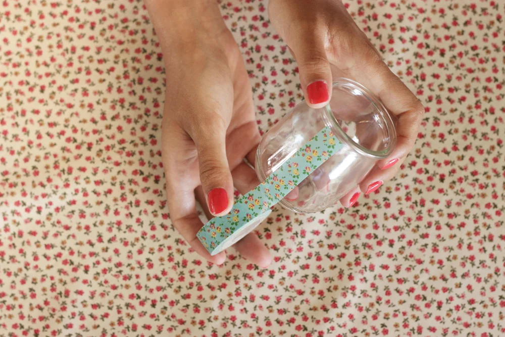 Mains aux ongles rouges décorant un photophore en verre avec un ruban floral sur un fond de tissu à petites fleurs.