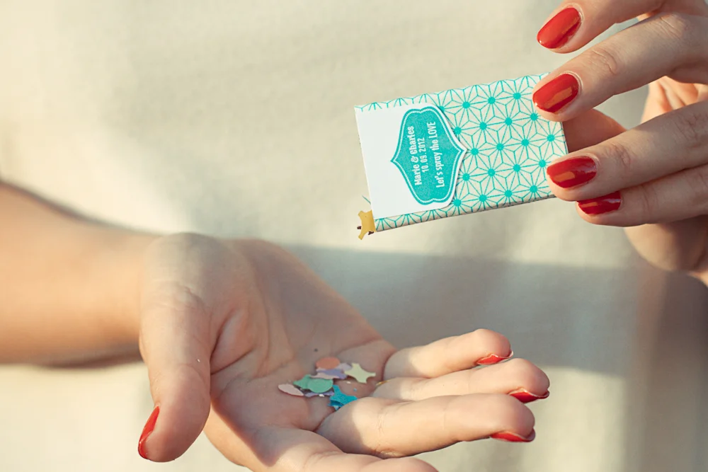 Une main verse des confettis colorés depuis un petit sachet turquoise personnalisé dans une autre main ouverte.