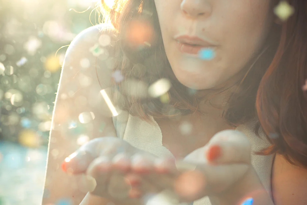 Gros plan lumineux d'une femme soufflant des paillettes colorées depuis ses mains dans une ambiance onirique.