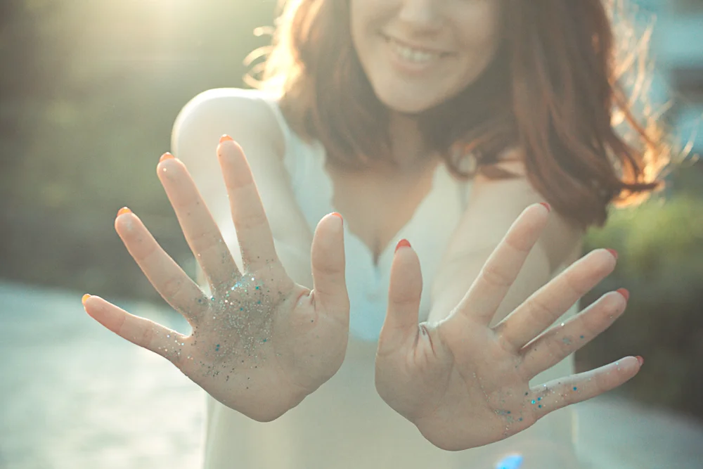 Gros plan sur les mains d'une femme souriante et lumineuse, parsemées de paillettes bleues et argentées.