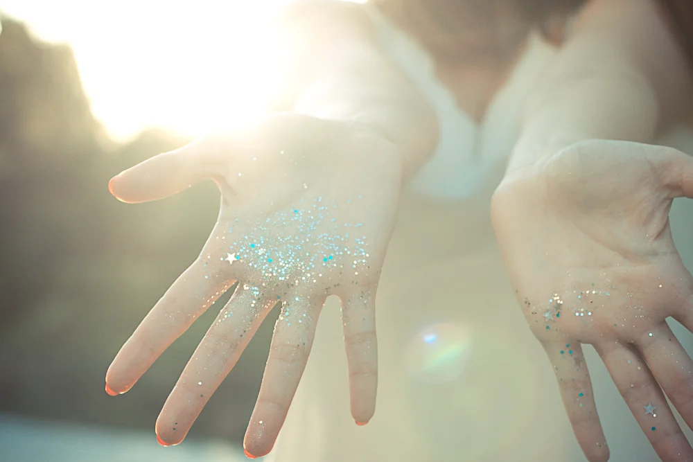 Gros plan lumineux sur des mains ouvertes parsemées de paillettes scintillantes dans une ambiance douce et vaporeuse.