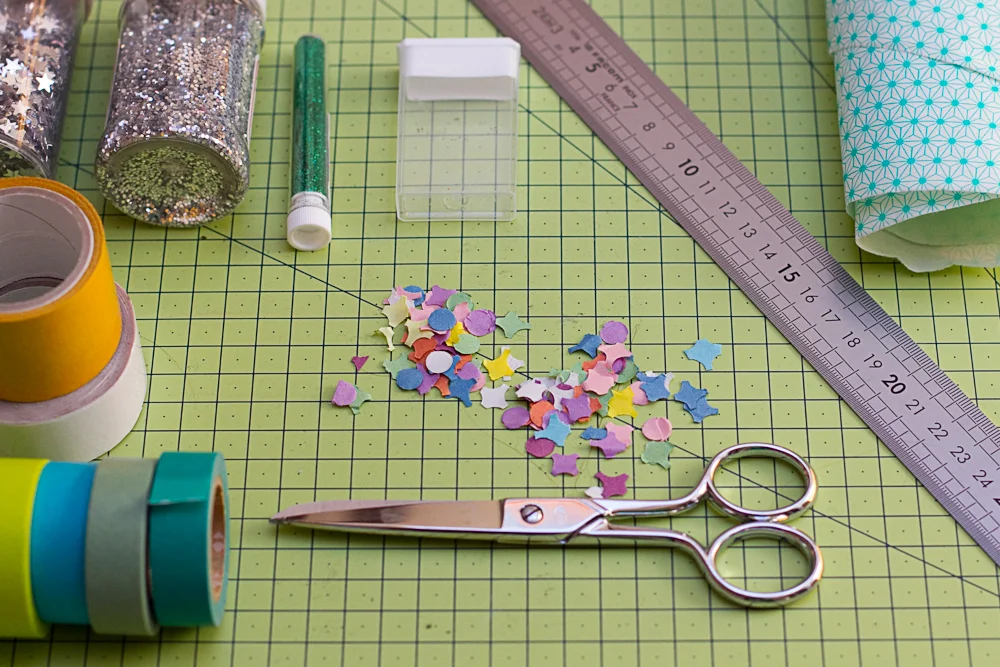 Fournitures de loisirs créatifs colorées, paillettes et ciseaux sur un tapis de découpe vert.