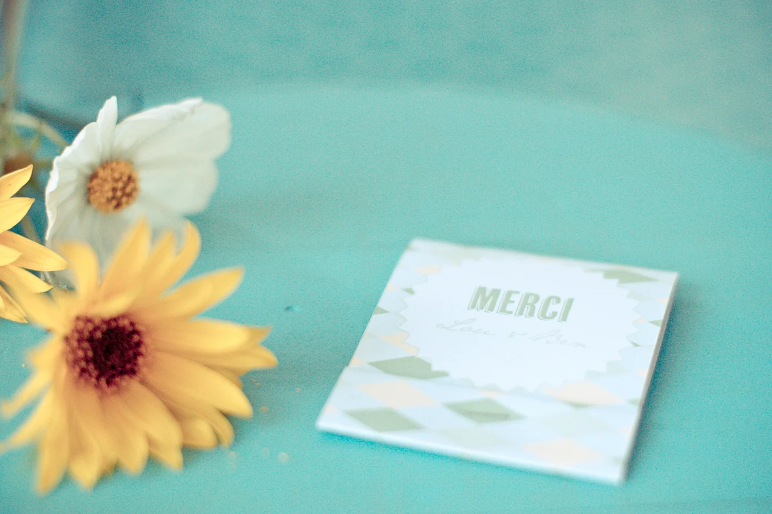 Carte de remerciement blanche et bleue posée à côté de fleurs jaunes et blanches sur un fond turquoise pastel.