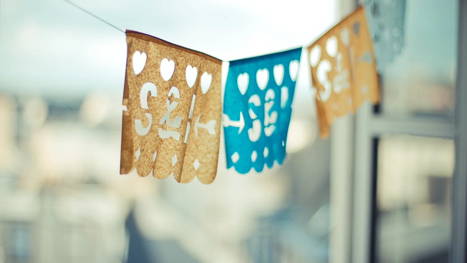 Guirlande de papel picado bleue et dorée suspendue devant une fenêtre, ambiance douce et lumineuse.