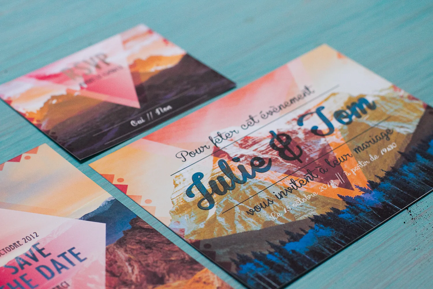 Faire-part de mariage colorés aux motifs montagneux et typographie cursive sur un fond bleu turquoise.