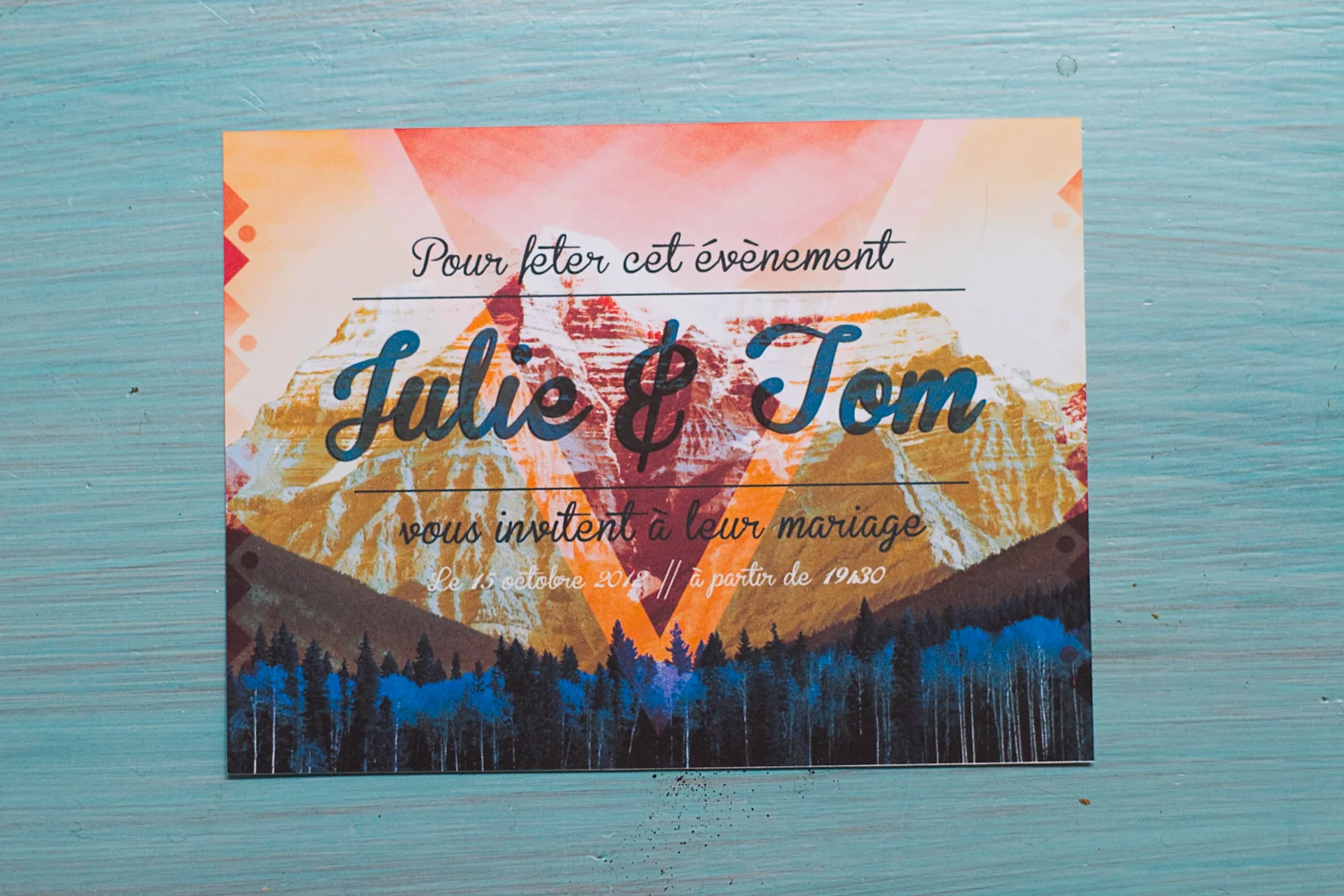Faire-part de mariage coloré avec paysage de montagnes et forêt, texte calligraphié sur fond bleu turquoise.