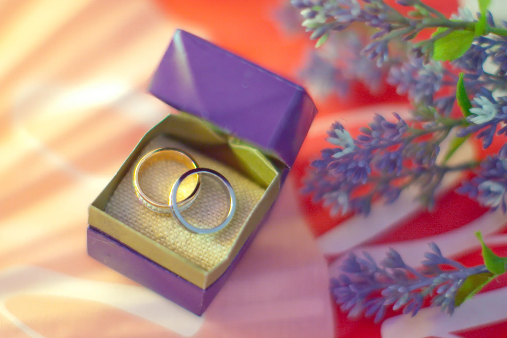 Deux alliances en or et argent posées dans un écrin violet ouvert, entourées de fleurs mauves sur un fond coloré.