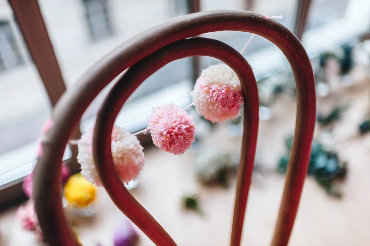 Guirlande de pompons roses et blancs suspendue à un dossier de chaise en métal, ambiance douce et festive.