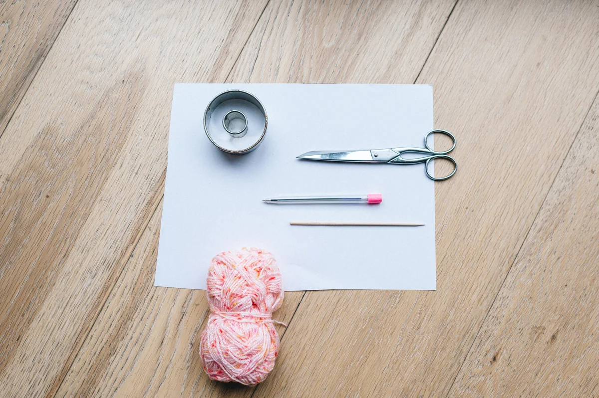 Matériel de couture composé de laine rose, ciseaux et accessoires posés sur une feuille blanche et un parquet clair.