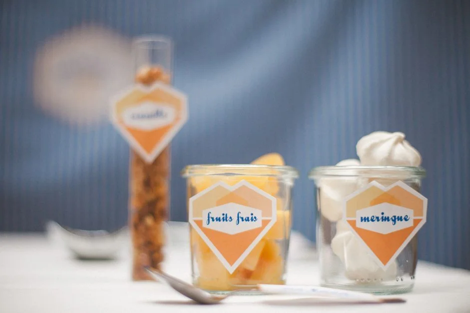 Trois contenants en verre avec étiquettes orange proposant des garnitures pour glaces sur un fond bleu.