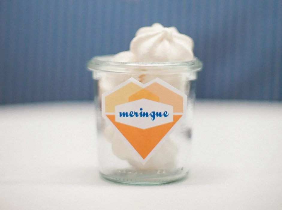 Meringues blanches dans un petit pot en verre avec une étiquette orange et blanche sur fond bleu et blanc.