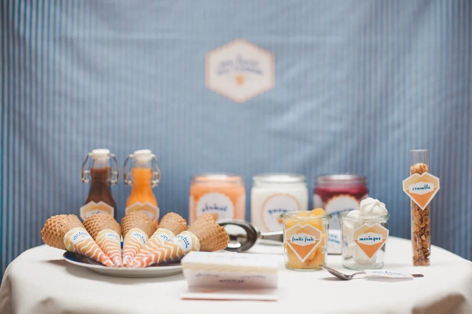 Bar à glaces DIY avec cornets, toppings et sauces colorées sur une table nappée de blanc devant un fond bleu.