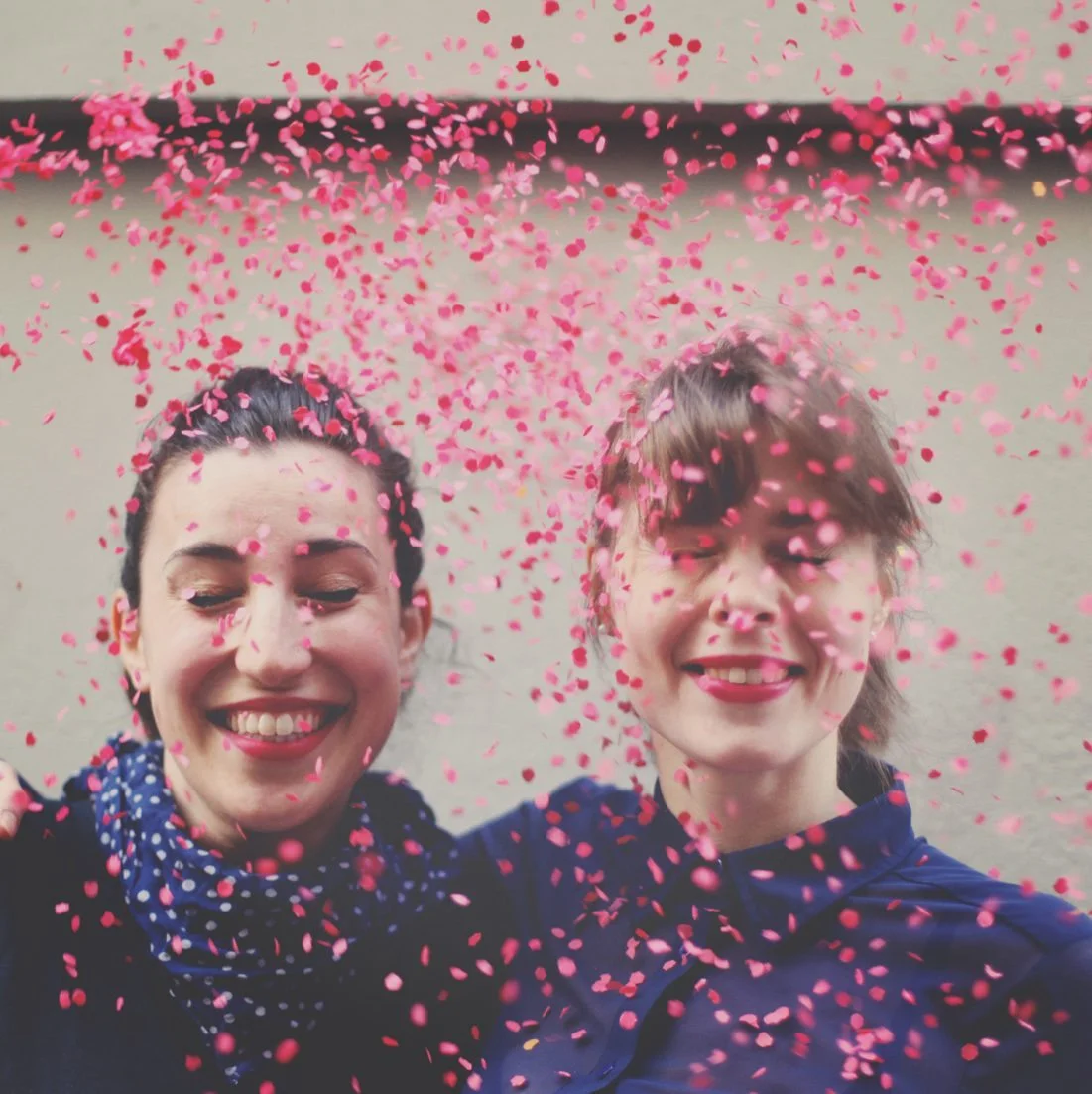 Deux femmes souriantes sous une pluie de confettis roses, dans une ambiance festive et joyeuse.