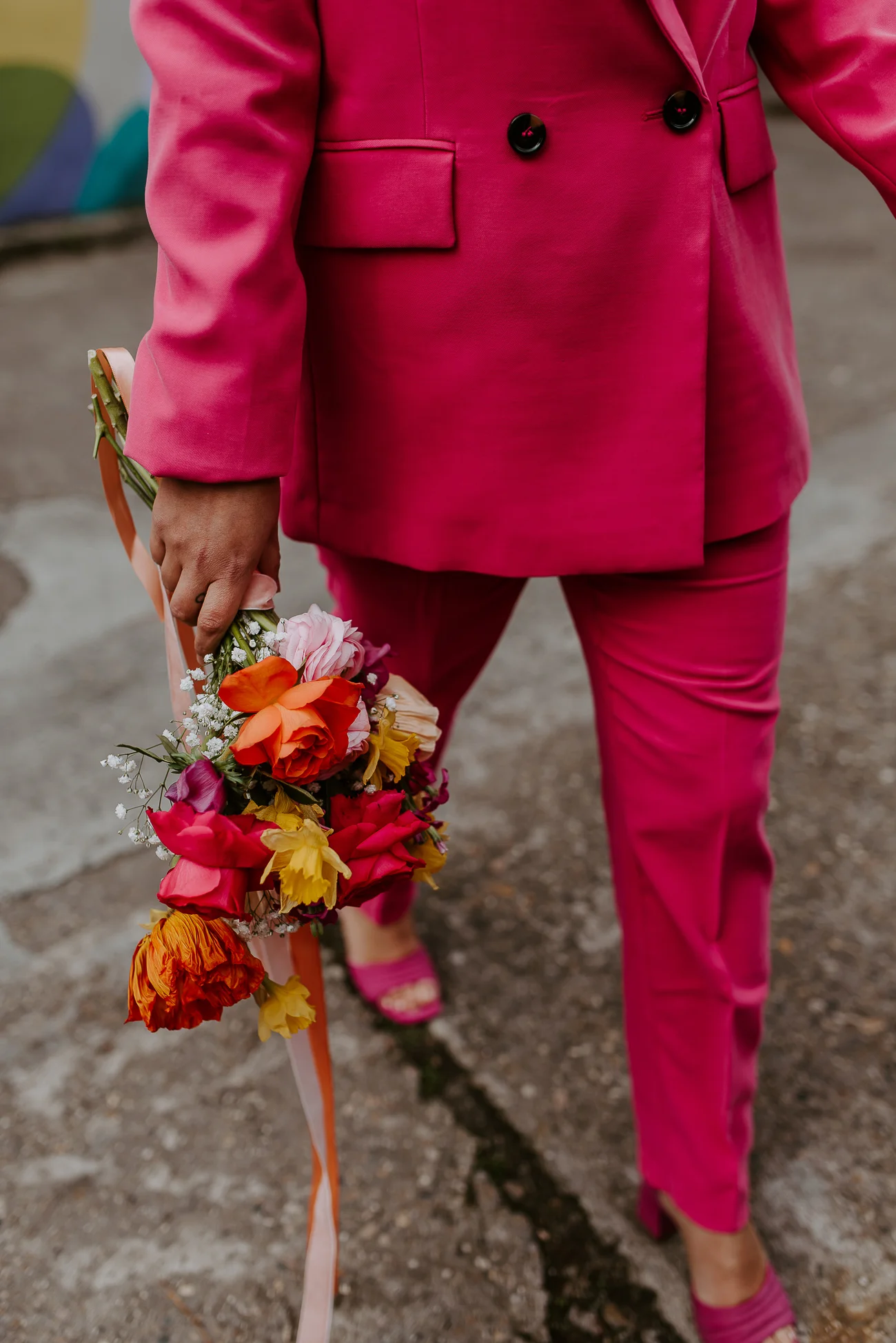 Un marié audacieux dans un costume fuchsia met en valeur son bouquet vibrante de fleurs rouges, oranges et jaunes,