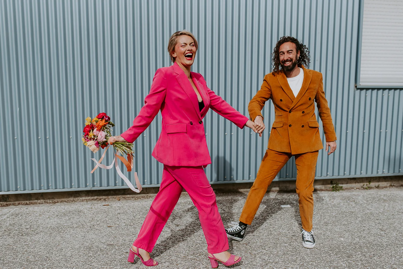 Une mariée en costume rose audacieux et son compagnon dans un costume rustique orange, marchant avec joie dans un décor
