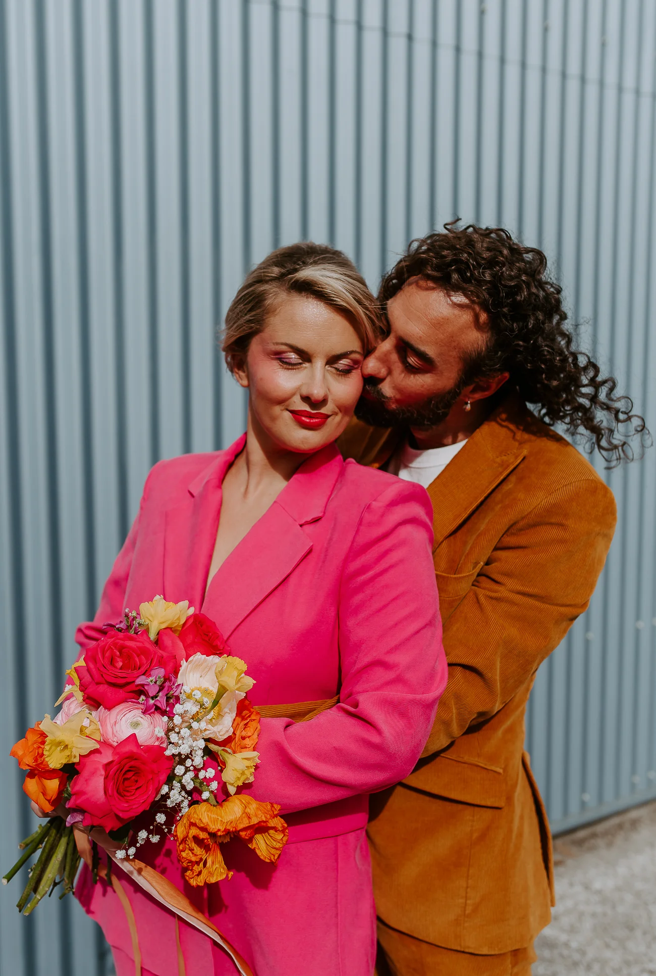 Une mariée en rose vif s'adonne à un moment tendre avec son compagnon, tenant un bouquet coloré de fleurs.