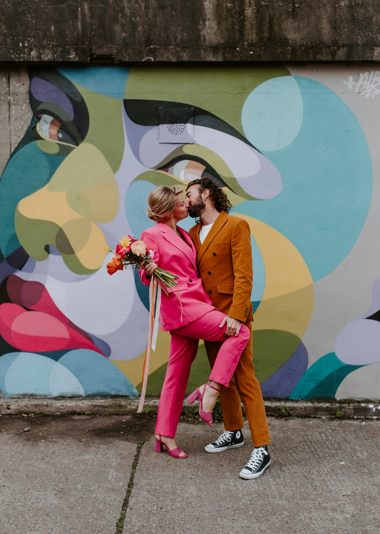 Une jeune mariée en costume rose tendre et un mari en costume rustique se baignant dans l'ambiance colorée d'un mur de