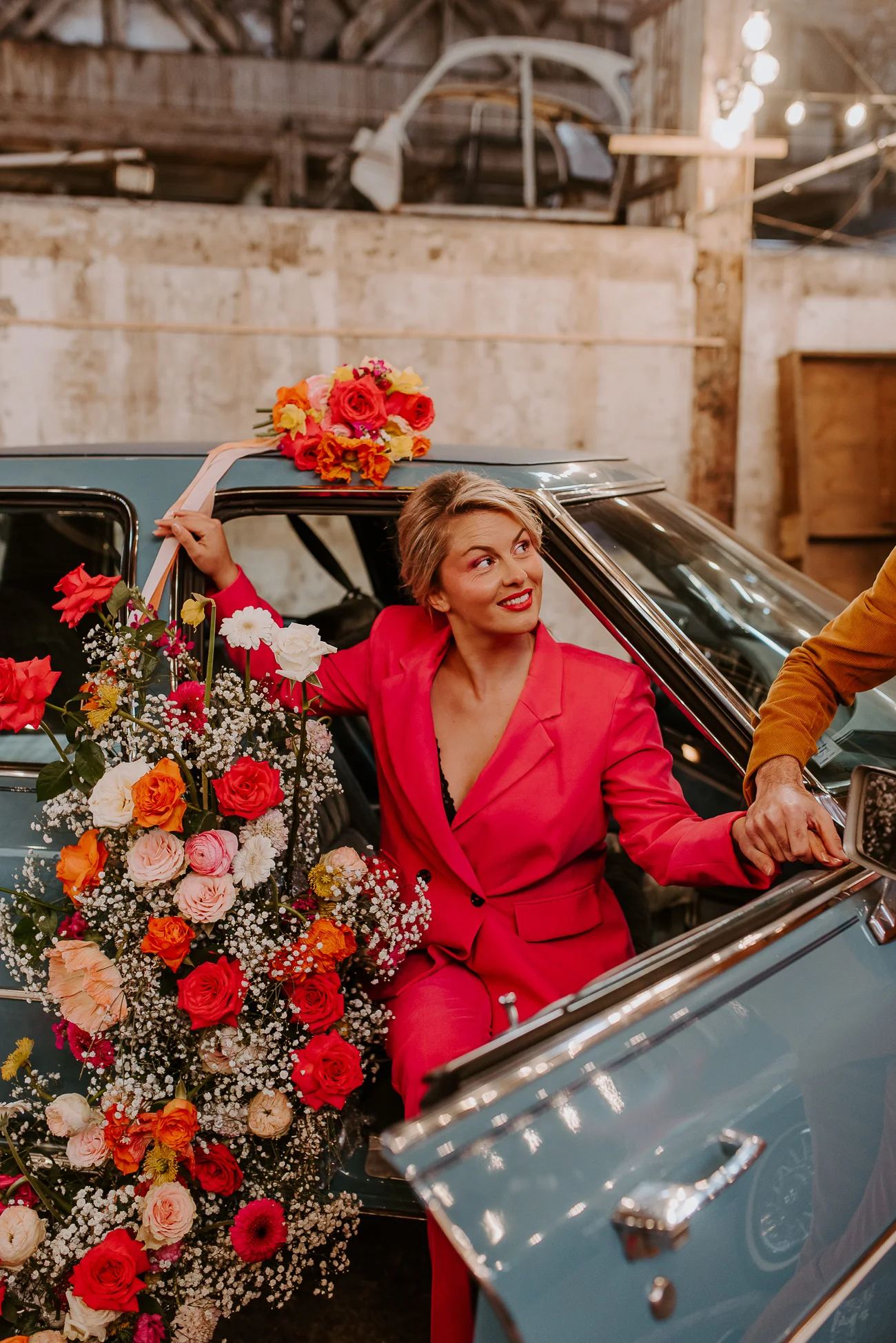Une jeune femme dans un costume rose saisissant, tenant des fleurs colorées, se détache contre un véhicule vintage dans