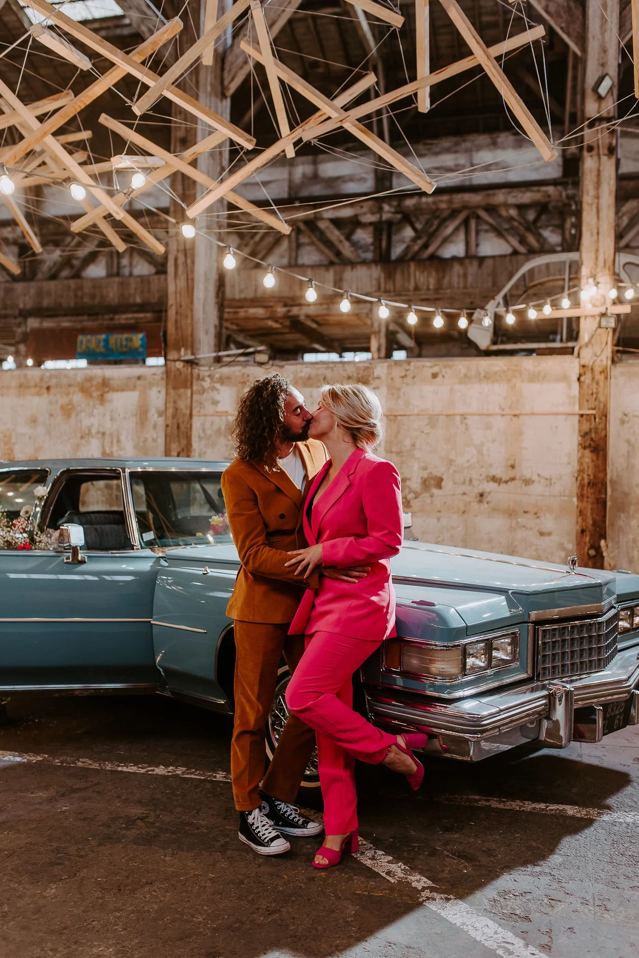 Un couple émerveillé dans des costumes vifs, partageant un baiser tendre devant un vintage bleu, plongé dans un décor