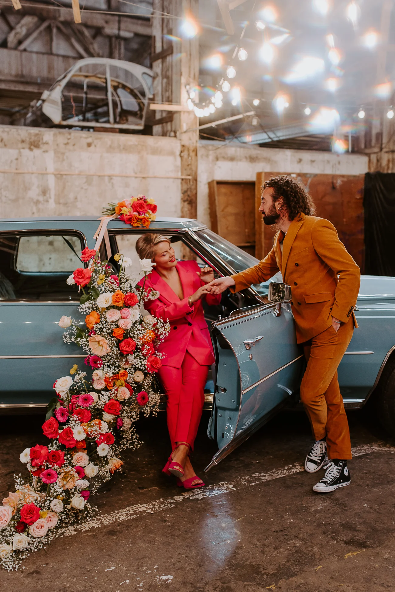 Un couple charmant debout devant une voiture bleue remplie de fleurs colorées, incarnant l'ambiance joyeuse d'un
