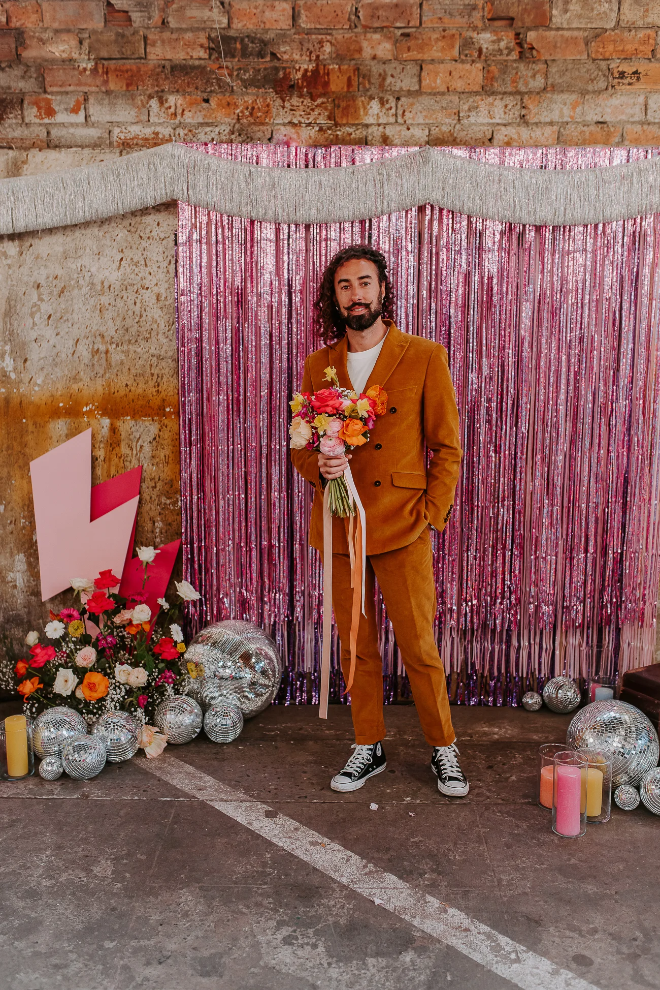 Un marié dans un costume orange flamboyant tient un bouquet coloré, entouré de décoration vibrante et lumineuse dans un