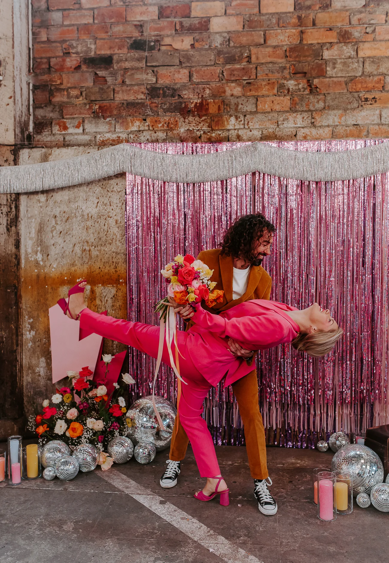 Un couple dans des costumes vibrants rose et orange dans une scène romantique avec des décorations en paillettes et des fleurs.
