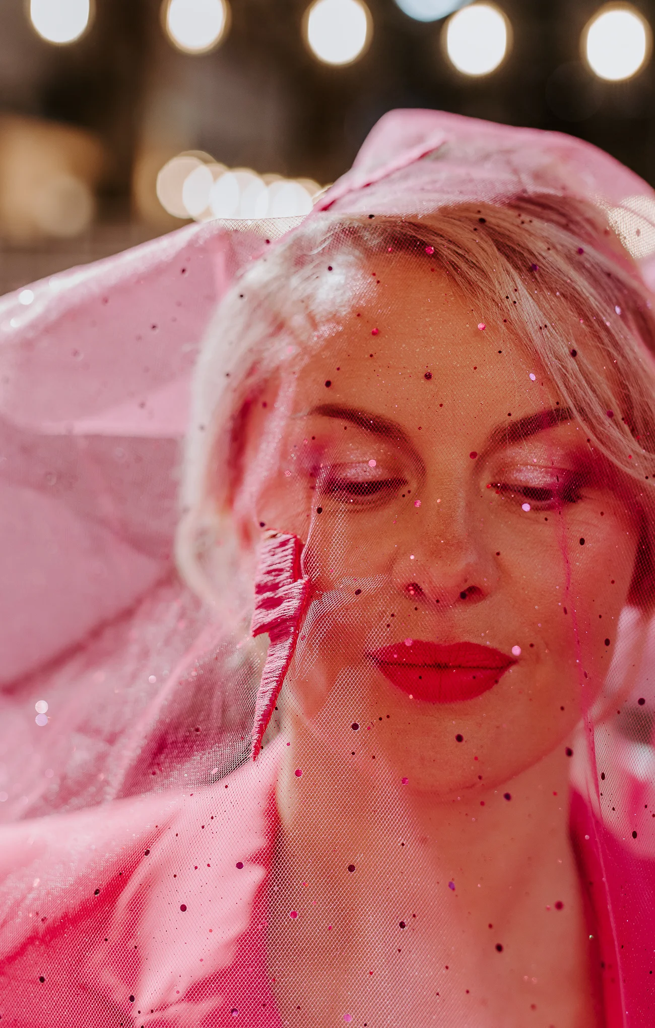 Une mariée dans une robe rose avec un voile, visage fermé les yeux, décoré de paillettes, avec un fond d'ambiance lumineuse.