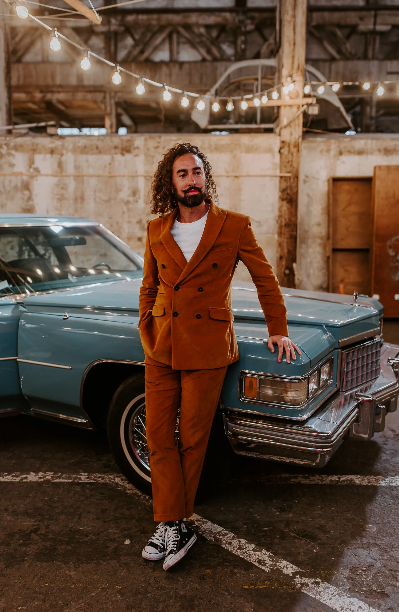 Un homme avec un style décontracté et raffiné se détache dans un garage vintage, une voiture bleue ancienne et des