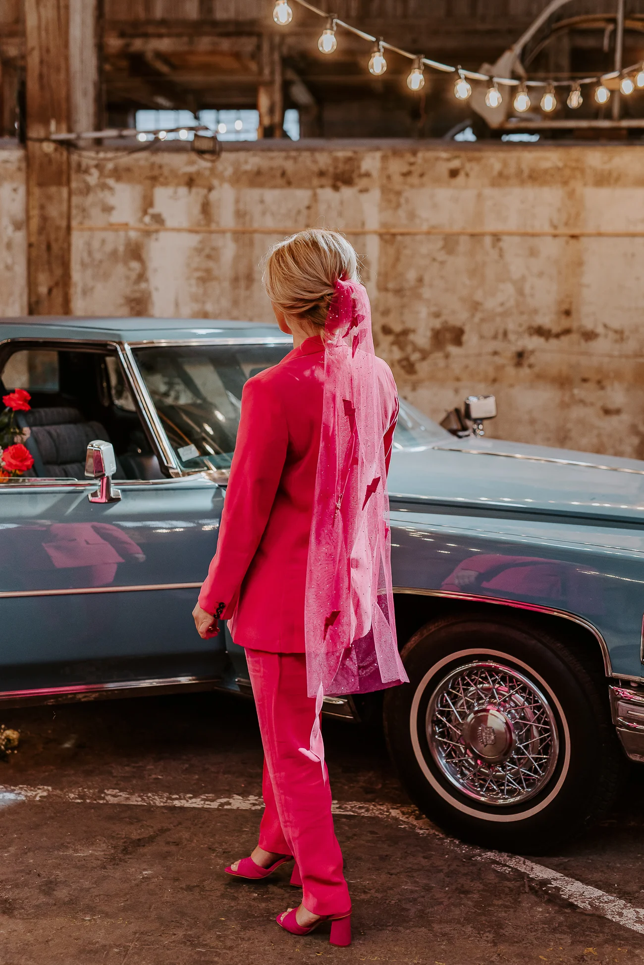 Une femme élégante dans un costume rose vif et un foulard pailleté, se dirigeant vers une voiture vintage bleue, dans