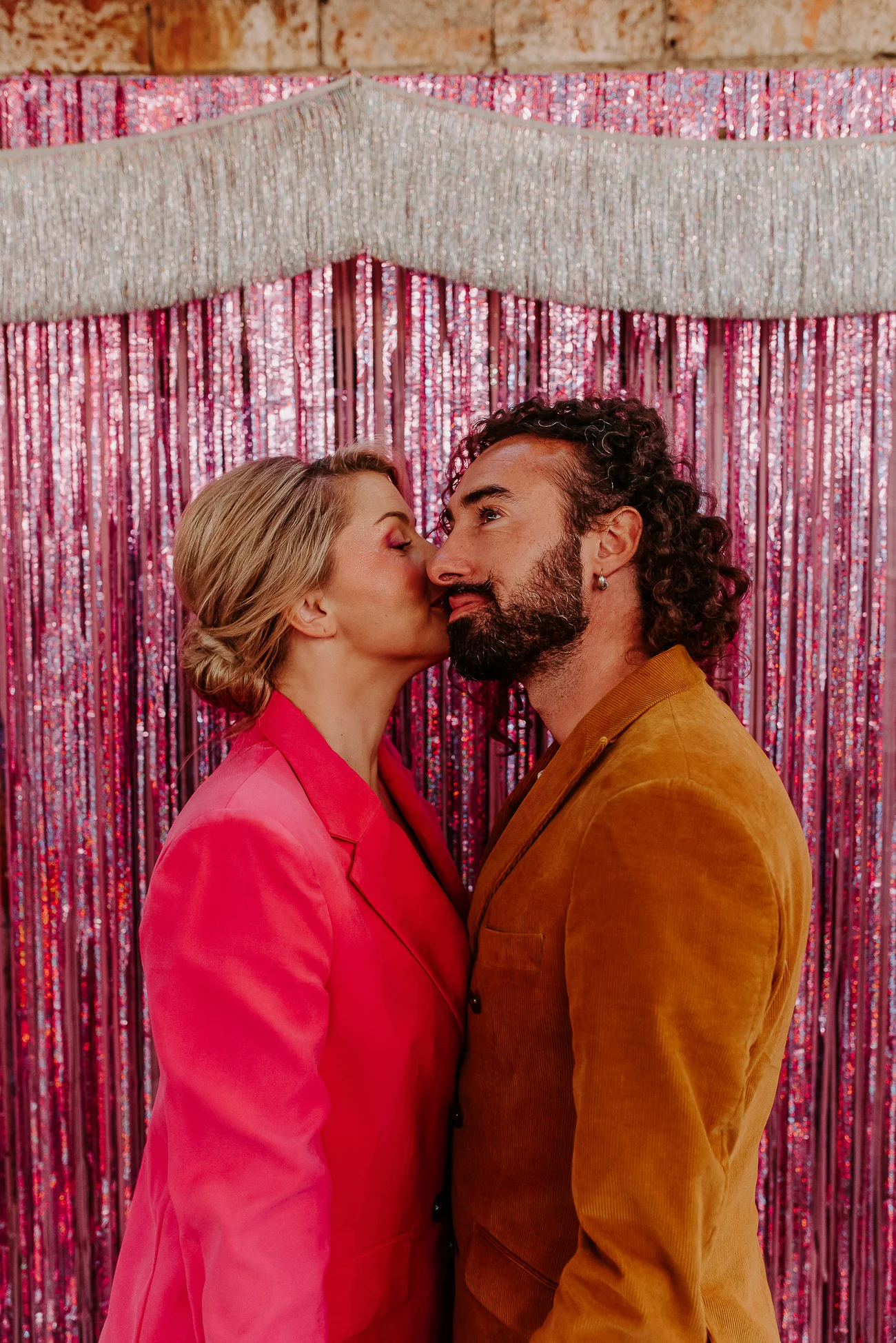 Un couple romantique partage un baiser tendre contre un fond de rideaux paillettes rose, incarnant l'ambiance énergique