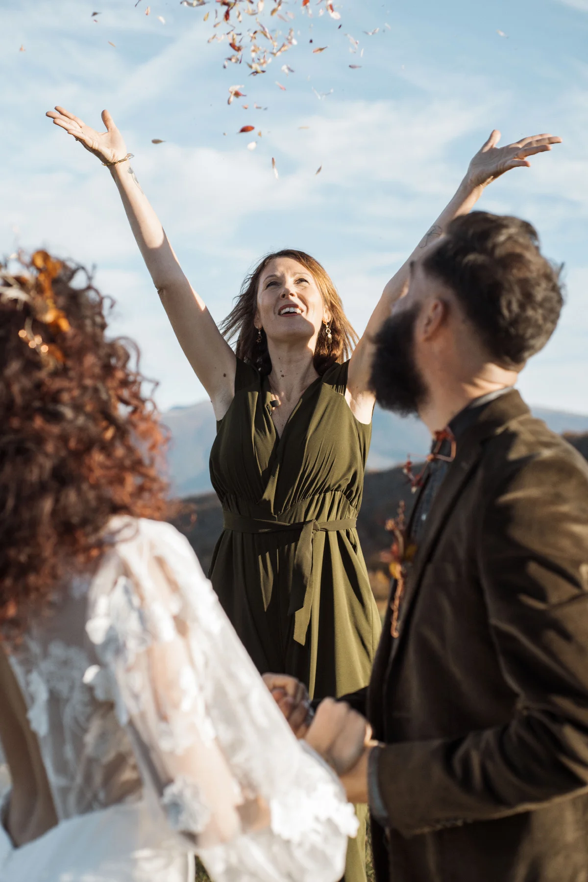 Une femme en robe vert olive lance des pétales en l'air lors d'un mariage folk en plein air.