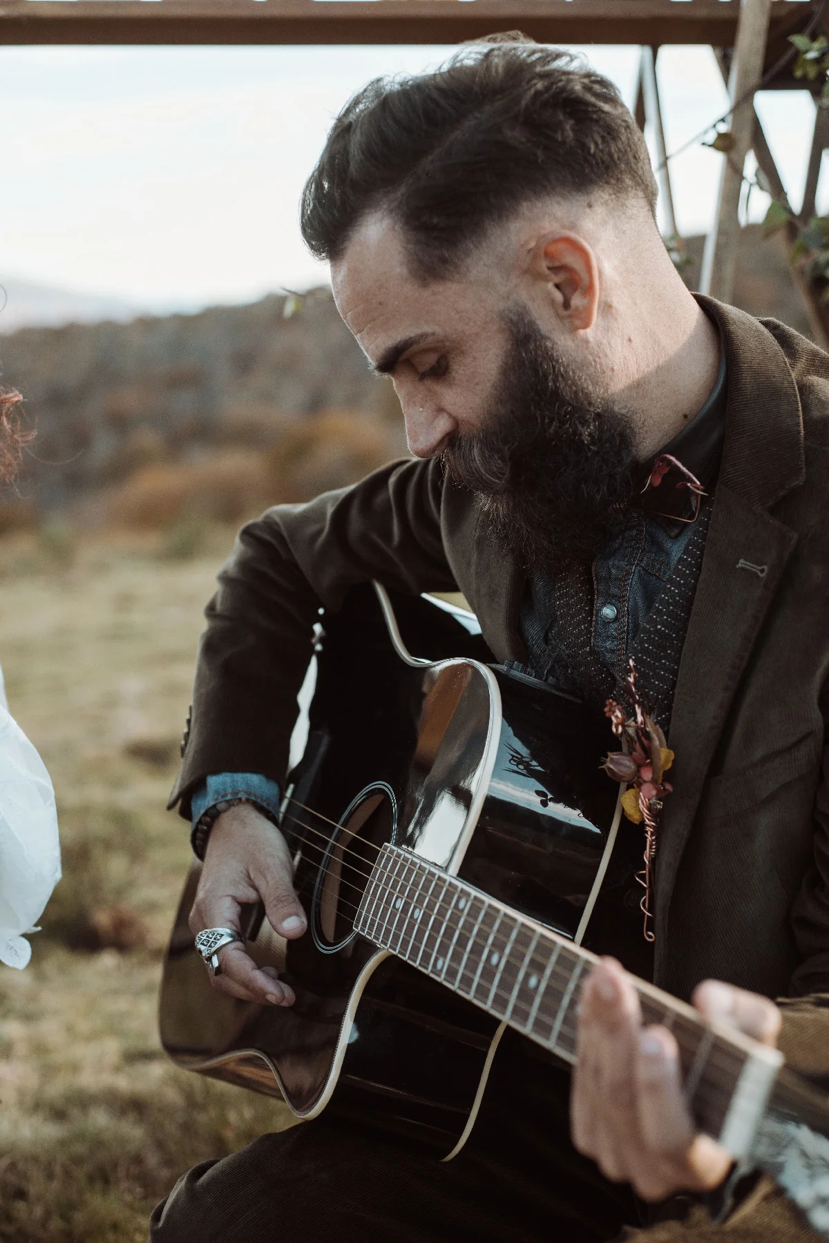 Homme barbu en costume marron jouant de la guitare acoustique lors d'un mariage folk en plein air.