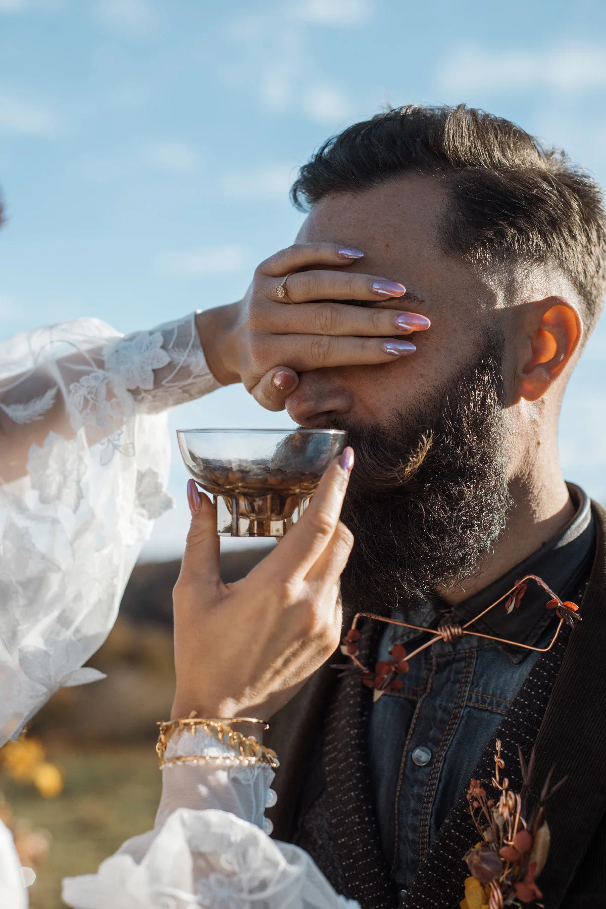 Une mariée en dentelle blanche cache les yeux de son époux barbu tenant un bol, dans une ambiance folk et naturelle.