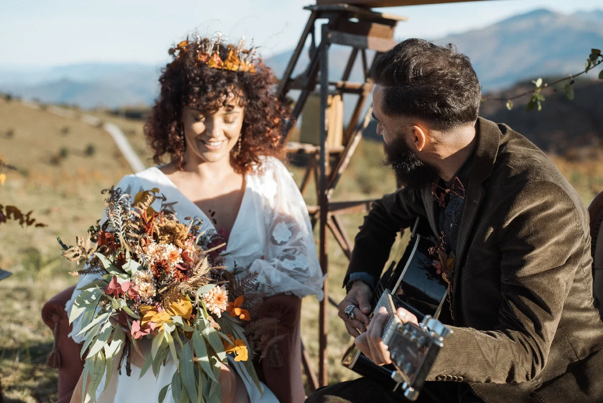 Mariée en robe blanche et couronne de fleurs souriant à un musicien à la guitare lors d'un mariage folk en plein air.