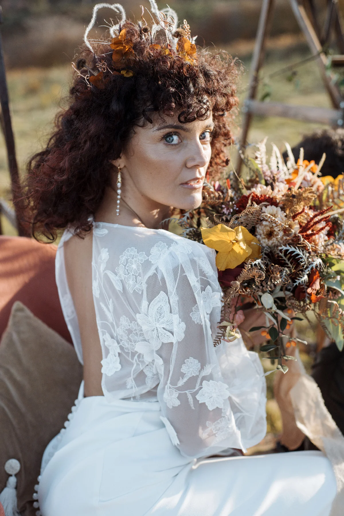 Mariée aux cheveux bouclés portant une robe blanche en dentelle et une couronne florale, tenant un bouquet champêtre.