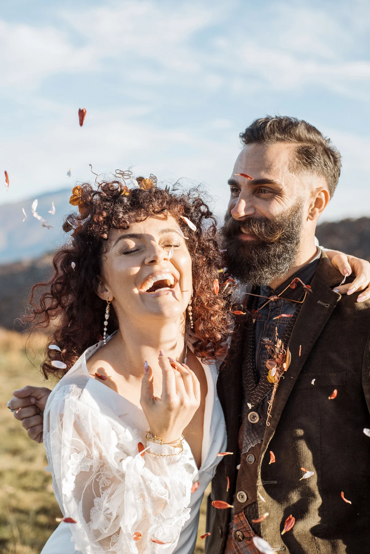 Couple riant sous une pluie de pétales, look folk et bohème, en extérieur avec des montagnes en arrière-plan.