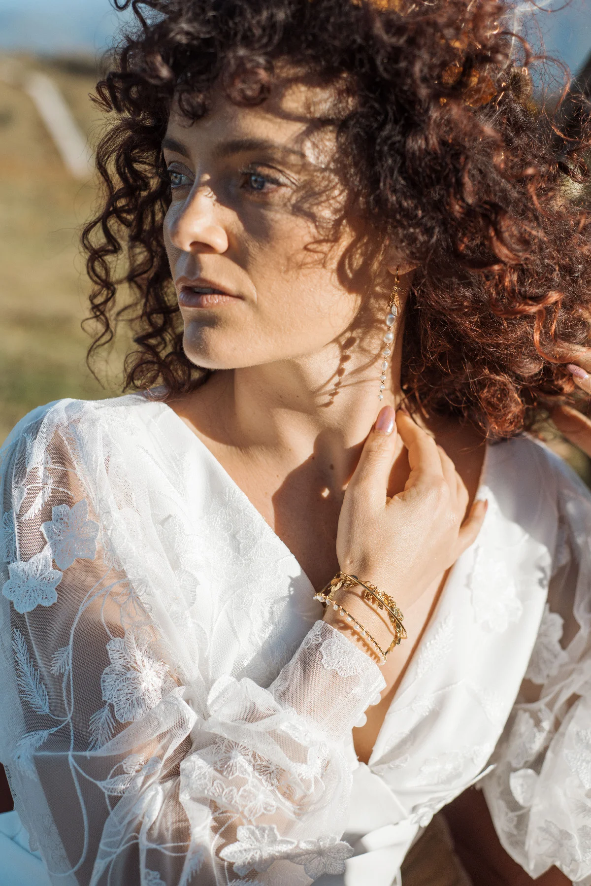 Mariée aux cheveux bouclés en robe blanche dentelle, posant en extérieur sous une lumière naturelle et chaleureuse.
