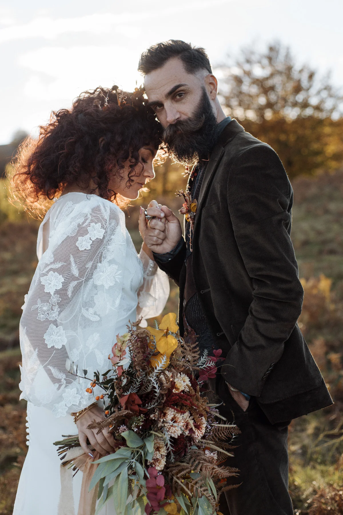 Couple en tenue folk, la mariée en dentelle blanche et le marié en costume brun, s'enlaçant dans un champ doré.