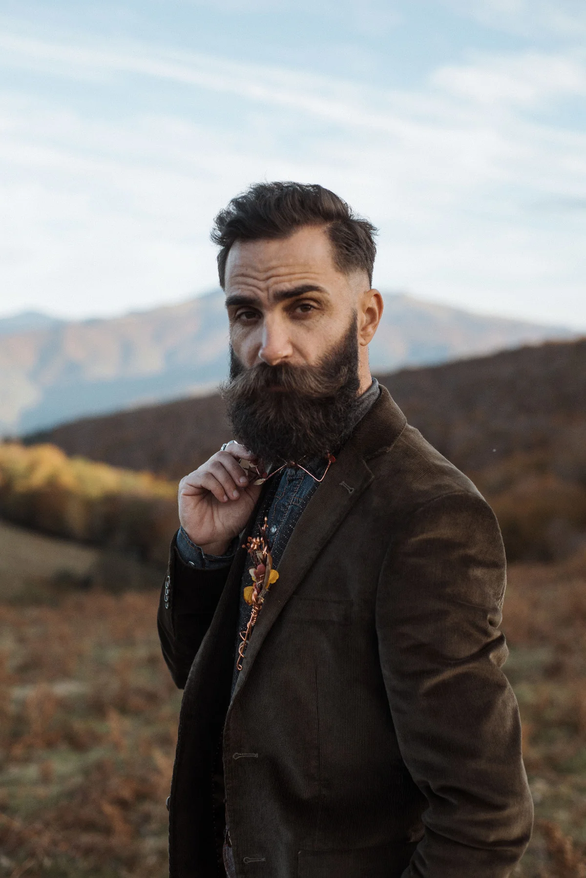 Homme barbu en costume marron et nœud papillon, posant dans un paysage naturel et montagneux aux tons automnaux.