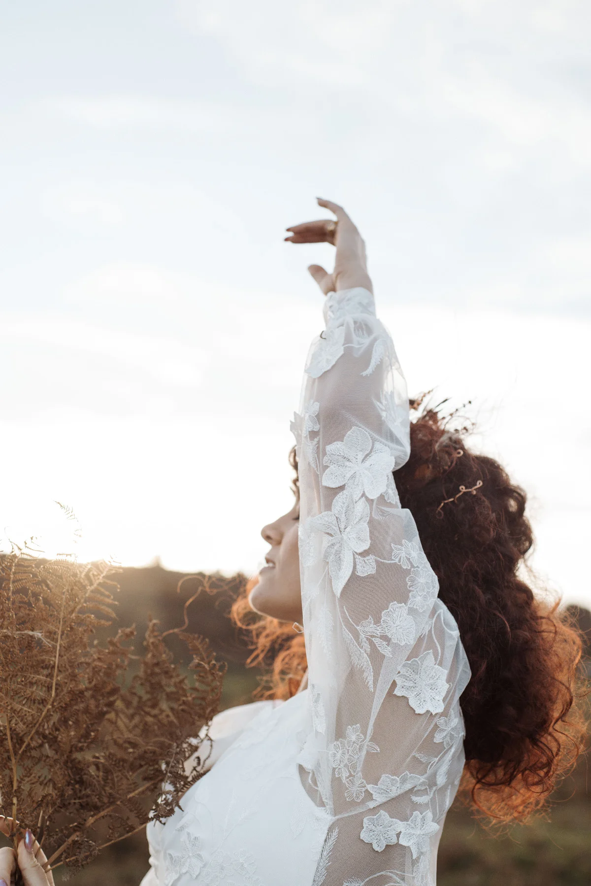 Mariée aux cheveux bouclés en robe blanche dentelée, levant le bras vers le ciel dans un paysage naturel et lumineux.