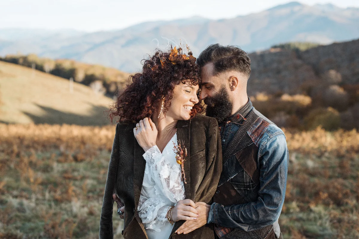 Couple complice en tenue folk et bohème s'enlaçant dans un paysage montagneux aux couleurs automnales.
