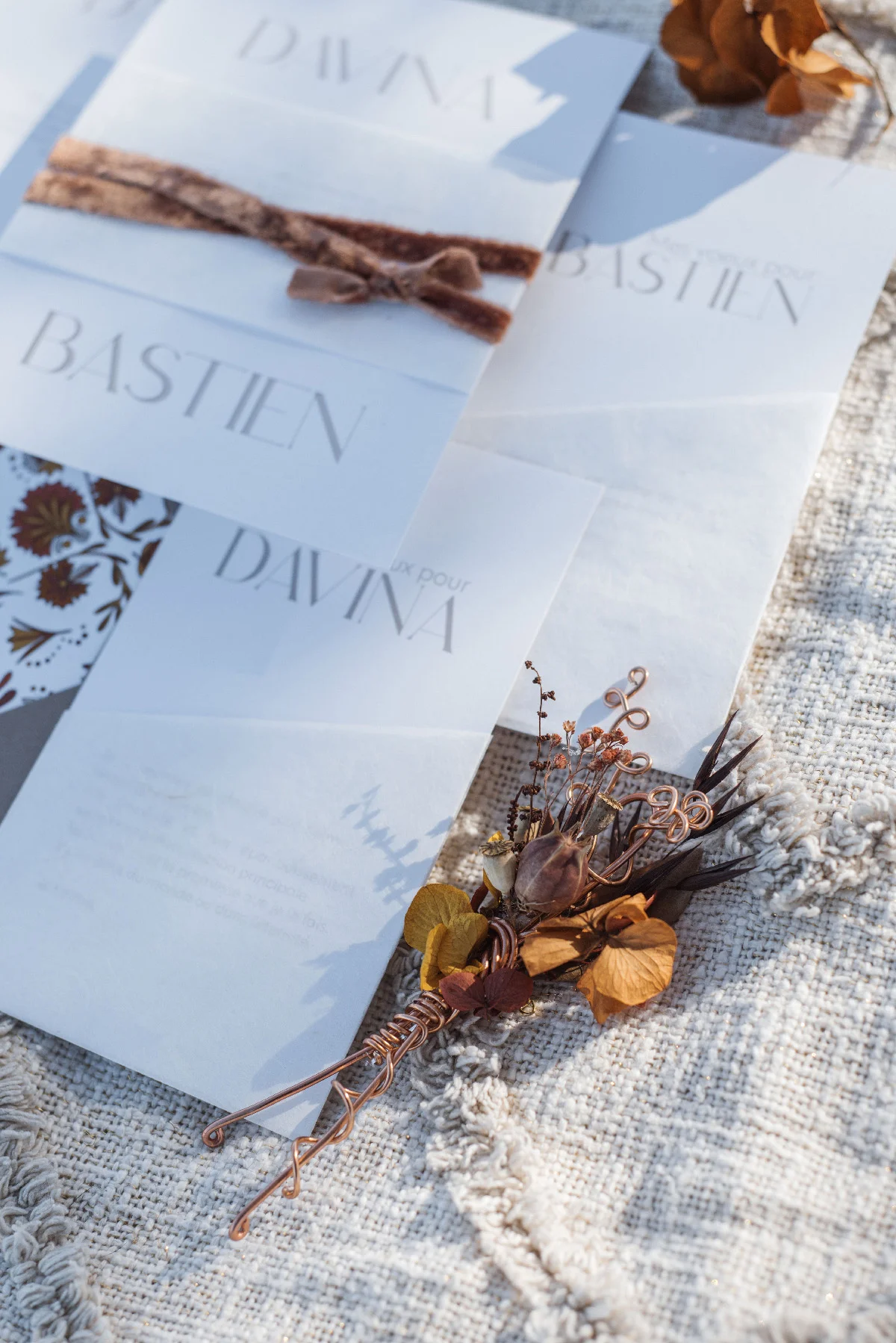 Papeterie blanche aux noms Davina et Bastien avec un petit bouquet séché aux tons terreux sur un tissu rustique.