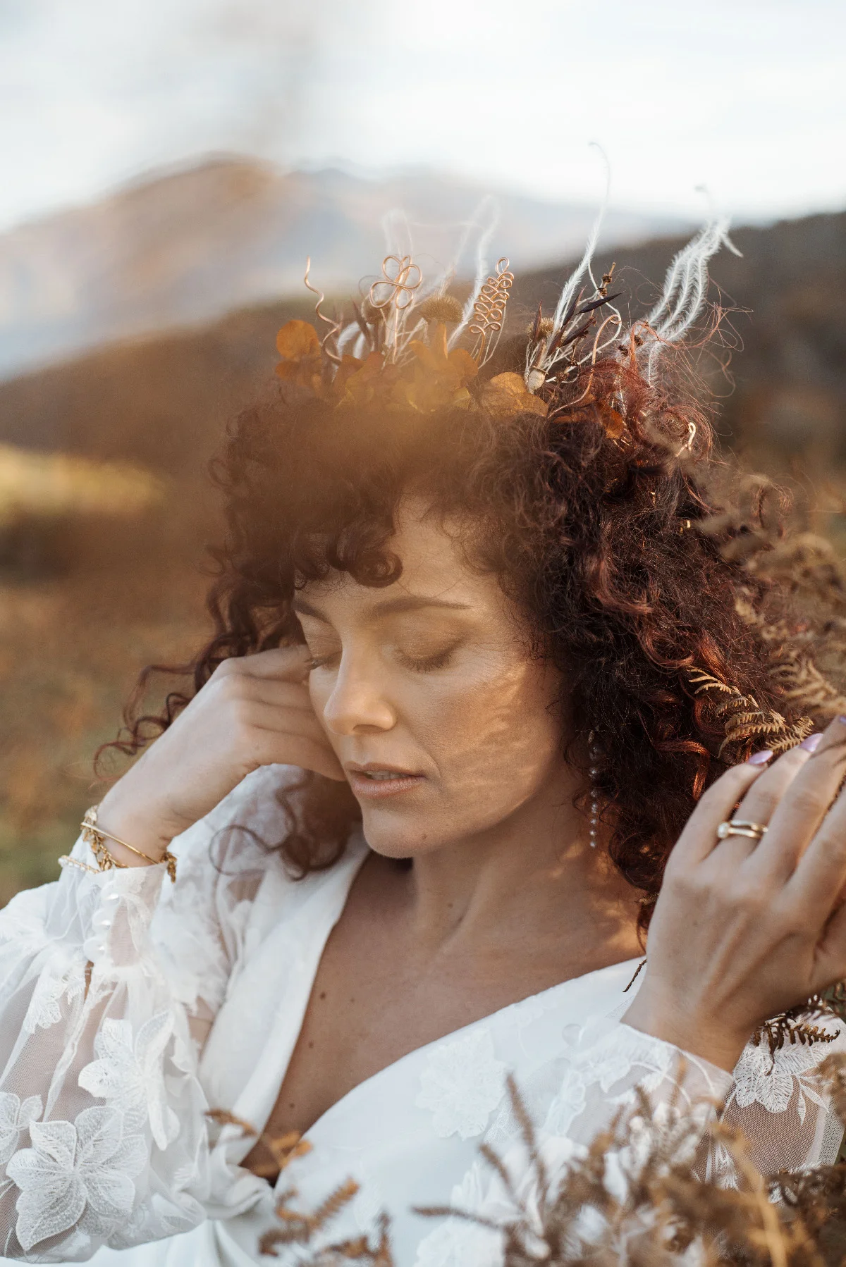 Femme aux cheveux bouclés portant une couronne végétale et une robe blanche en dentelle dans un paysage naturel doré.