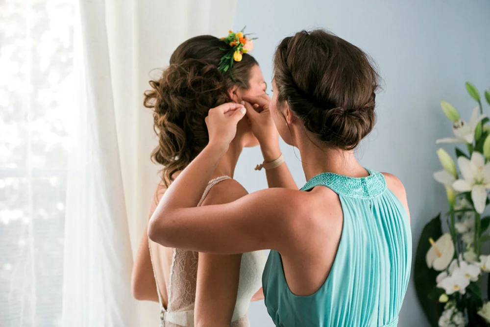 Une demoiselle d'honneur en robe turquoise aide la mariée à mettre ses boucles d'oreilles dans une ambiance douce.