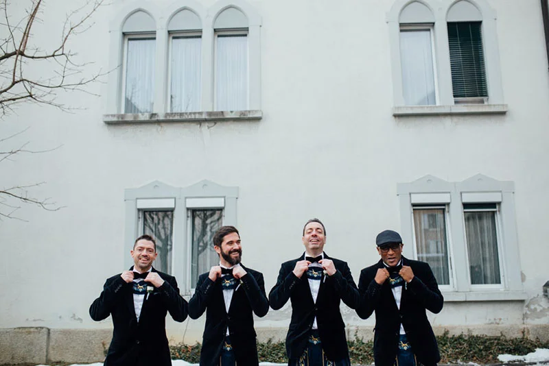 Quatre hommes souriants en costumes noirs et kilts traditionnels posent devant un bâtiment blanc.