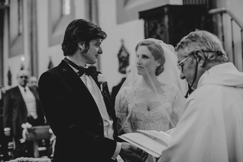 Photo noir et blanc d'un couple, Victoire et Xavier, s'échangeant des vœux devant un prêtre dans une église.