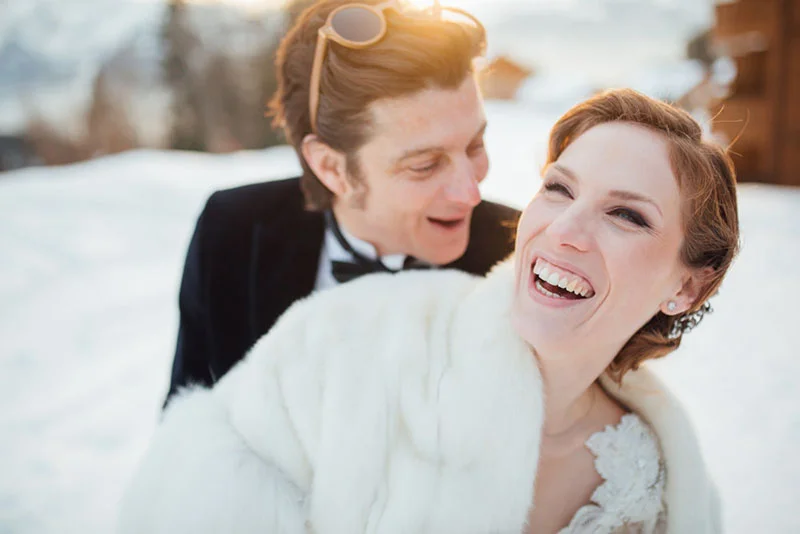 Couple riant dans la neige, la mariée en étole blanche et le marié en smoking, ambiance hivernale et romantique.