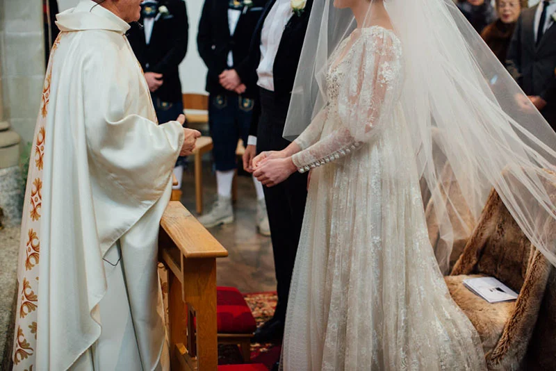 Une mariée en robe de dentelle blanche et voile long face à un prêtre lors d'une cérémonie religieuse solennelle.