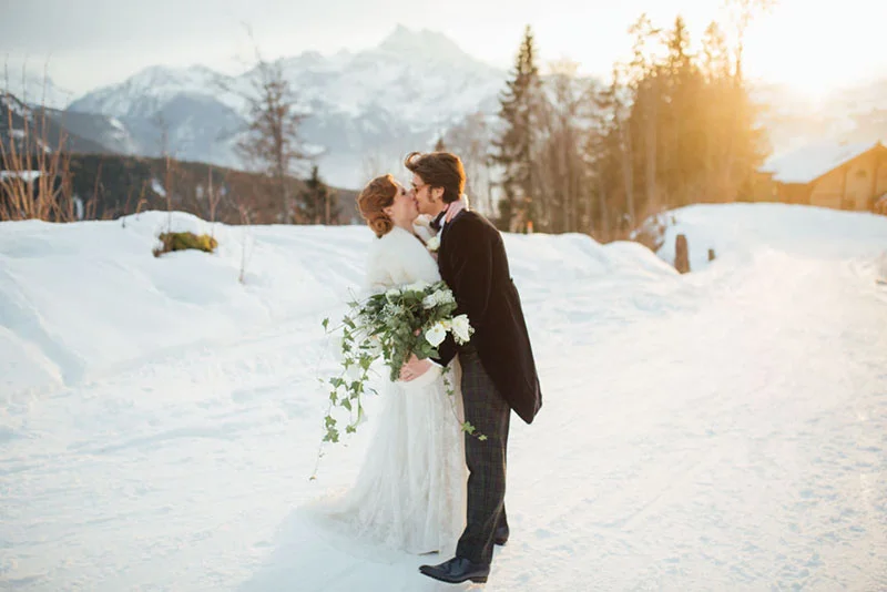 Jeunes mariés s'embrassant dans un paysage enneigé de montagne au coucher du soleil, ambiance romantique et hivernale.