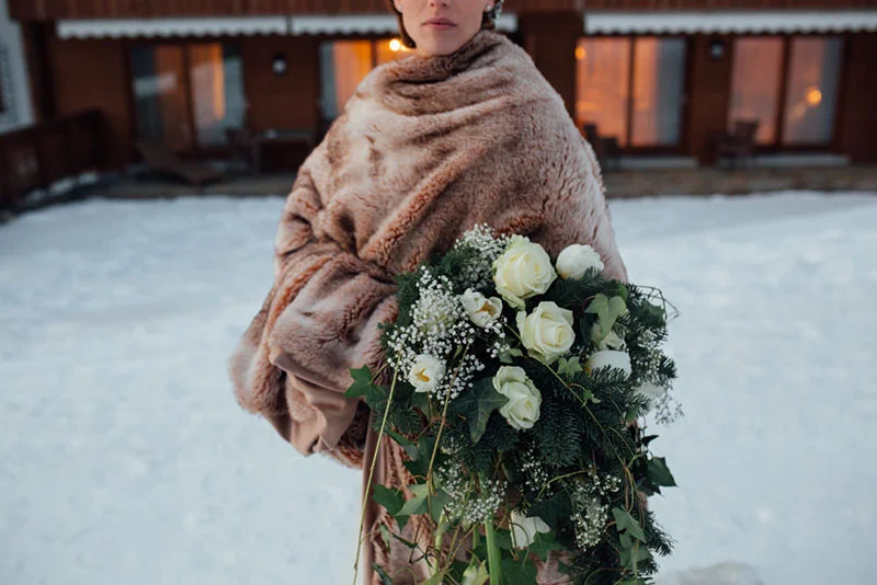 Mariée enveloppée dans une étole en fourrure beige tenant un bouquet de roses blanches sur un fond de neige.